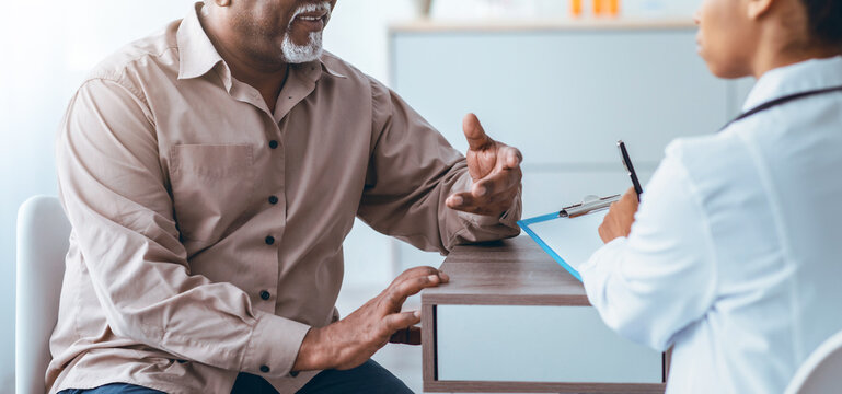 A man speaks with a healthcare provider in a medical office. They discuss health matters while the provider takes notes. The setting is bright and organized, focusing on patient care.