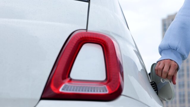 Close up of female hands opens vehicle charge port and putting charger cable into electric car. Woman driver charges electro vehicle at charging station outdoor. EV concept, eco car.