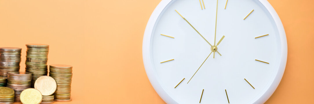 Concept of wealth accumulation and time with gold coin stacks and white wall clock on orange background.