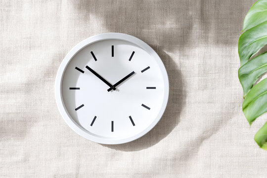 Minimalist white wall clock on beige linen texture background with green tropical leaf.