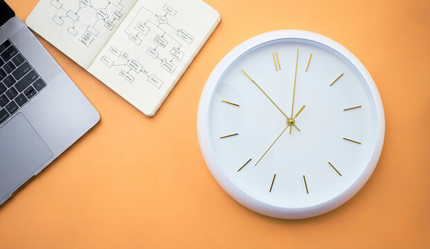 Business planning and time management concept with laptop, notebook with flowchart and clock on orange background