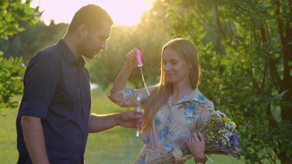 Fototapeta premium Close up of cute couple in love together outside. Man and woman with flowers standing in park and blowing soap bubbles. Love and romantic. Nature. Boyfriend and girlfriend