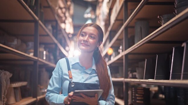 Focused young Asian woman in a blue apron checks a tablet in a long warehouse aisle dramatically lit from behind, creating a striking cinematic atmosphere.
