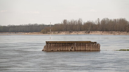 Zdjęcie przedstawia szeroką rzekę w surowym, wczesnowiosennym lub zimowym krajobrazie. Na środku nurtu widać rdzewiejącą, metalową konstrukcję barki, a w oddali piaszczysty brzeg. © tomo81