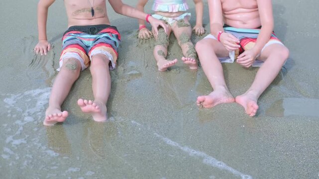 three children sibling sitting on sandy beach with feet in shallow ocean water, enjoying waves and summer fun together during family vacation, kids playing seaside with sand and water, childhood joy