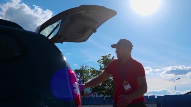 Young courier driver in cap unloading cardboard boxes from car while standing outdoor. Parcel delivery, service, business occupation concept. Real time