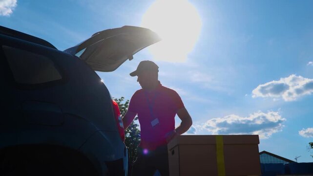 Young courier carefully loading a lot of cardboard boxes in the car in the trunk of a minivan during daytime. Parcel delivery, service concept. Real time