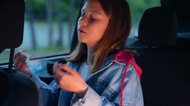 Girl sitting in the back seat in a car and fastening her seat belt, using a protective mask. Safety, transportation, healthy, childhood concept. Real time