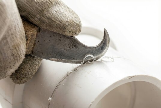 Plumber using a curved hook knife tool to deburr the cut edge of a white PVC plastic pipe