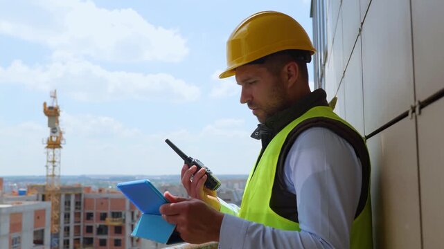 Male worker foreman wearing vest with helmet and using walkie talkie while holding tablet and inspecting housing estate at construction site. Real time video