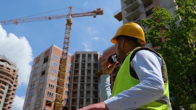 Back view of engineer in safety vest with helmet holding walkie talkie while standing at construction site. Inspector builder talking to colleagues on the radio. Real time video