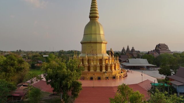 Breathtaking wide-angle drone footage of the golden Maha Chedi Sri Sarakham temple during a vibrant tropical sunset in Northeast Thailand.
