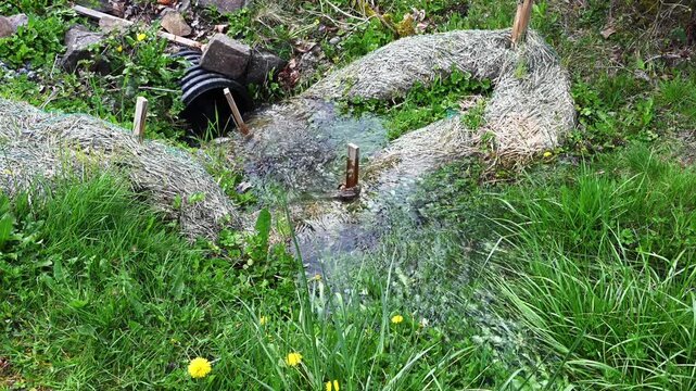 Water running in a grass filled ditch over a straw wattle and into black plastic stormwater system pipe, straw to catch debris and sediment to keep system clean
