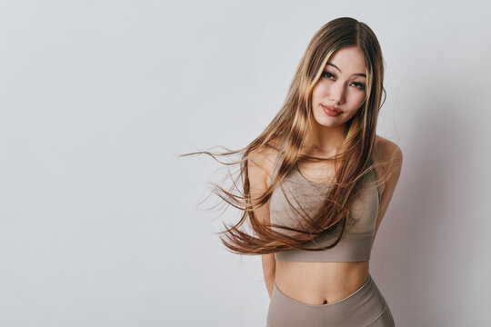 Young woman posing in beige sportswear with long hair flowing, standing against a light neutral background. Fitness, beauty, and casual lifestyle with natural makeup and confident expression.