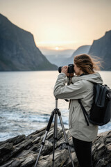 Fototapeta premium Woman photographing fjord at sunset with tripod on rocky shoreline and backpack