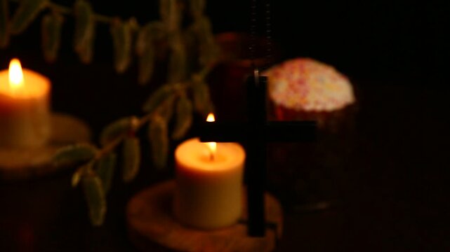 A hanging wooden black cross sways in front of the Easter candles flames near Easter cake on a dark blur background placed. No people