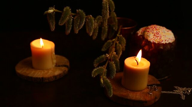 Two bright Easter burning candles with Easter cake on dark black background on natural wooden candlesticks, silver holy cross, willow branch on a table. Closeup warm concept, Easter holiday. No people