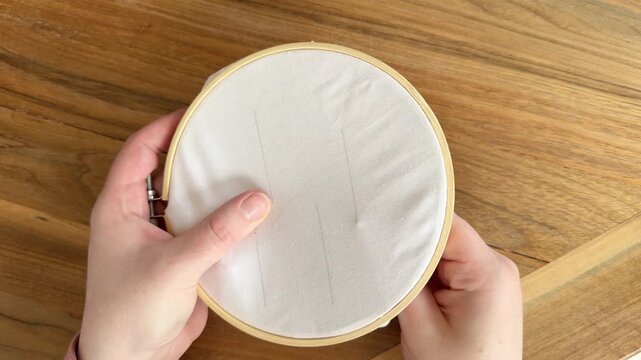 Female Caucasian hands pulling purple thread through fabric in an embroidery hoop, beginning a stitch on white cloth. Close-up 4K sewing and embroidery process on wooden table.
