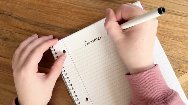 Female Caucasian hands writing &ldquo;Summer Budget&rdquo; on lined notebook paper, planning finances and organizing expenses. Close-up 4K writing process on wooden table.
