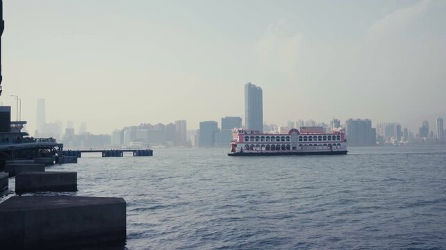 Static wide shot of Star Ferry crossingand arriving at pier at Victoria Harbour from Hong Kong Island side pier, ornate red and white vessel with Kowloon skyline and ICC tower visible in background.