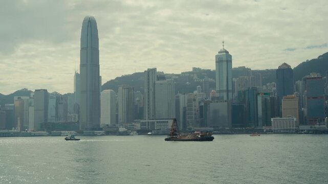 Static wide shot of dredging barge with crane and tugboat crossing Victoria Harbour, Two IFC tower and Hong Kong Island financial district skyline visible behind, overcast light, Hong Kong.