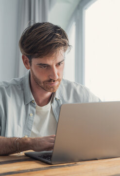 A young man concentrates on his laptop at a bright home office desk, typing and working remotely. Casual, modern workspace conveys productivity, focus and professional freelance or remote work