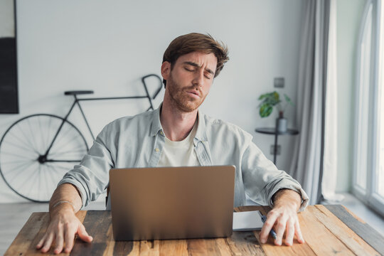 A young man sits at a wooden table with a laptop, eyes closed and head tilted back, expressing fatigue, stress and burnout from remote work in a modern, casual home office environment