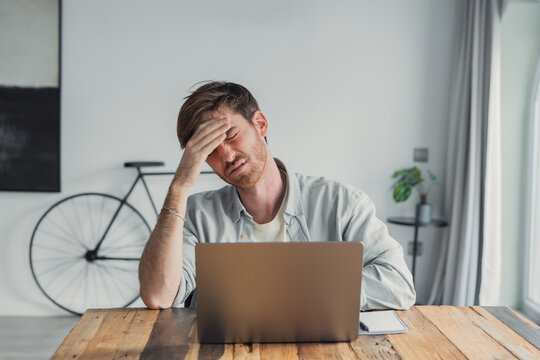 Young man at a wooden table works from home on a laptop, holding his head in frustration. Image conveys stress, tiredness, remote work challenges, concentration problems and low energy in a modern 