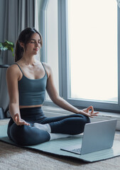 A young woman sits cross-legged on a mat by a bright window, meditating during an online yoga and...