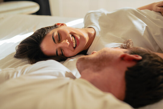 A young couple lies in bed laughing and sharing a tender moment, enjoying a relaxed morning at home with warm natural light that conveys intimacy, joy, connection and everyday romance.