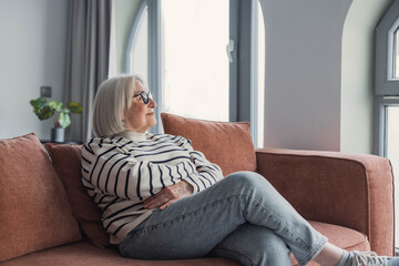 Obraz premium Elderly woman in a striped sweater sits comfortably on a sofa in a bright living room, gazing out the window with a calm, reflective expression that suggests relaxation and contentment.