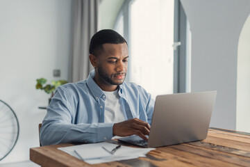 A young man concentrates while typing on a laptop at a wooden table in a bright home office,...
