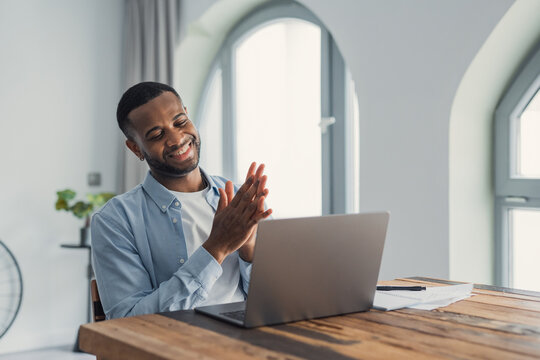 A smiling man applauds in front of his laptop, celebrating a successful remote work moment in a bright, modern home office. Papers and a notebook sit on the wooden table beside him.