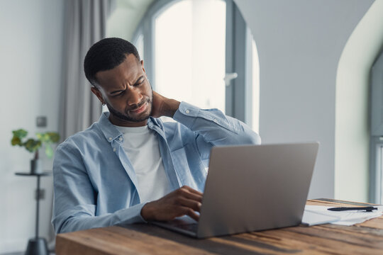 Young man at a wooden desk in a home office rubs his neck while working on a laptop, showing discomfort from long screen time, poor posture and remote work strain. Documents and pen nearby.