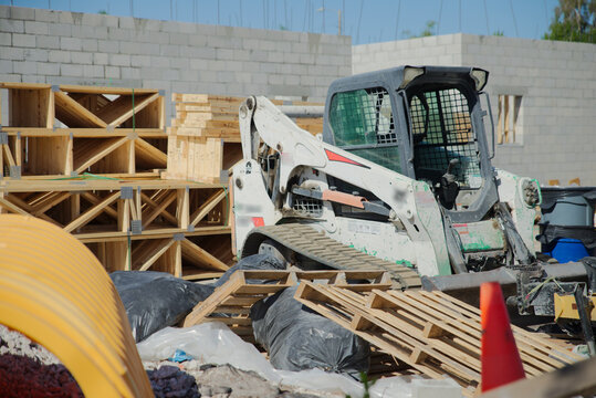 Skid steer loader parked among wooden pallets, plastic-wrapped debris and concrete block walls at a busy residential construction site, conveying industry, work in progress and on-site equipment.
