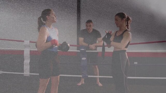 Two women boxers tightening black gloves, wraps after coach gesturing, preparing to spar