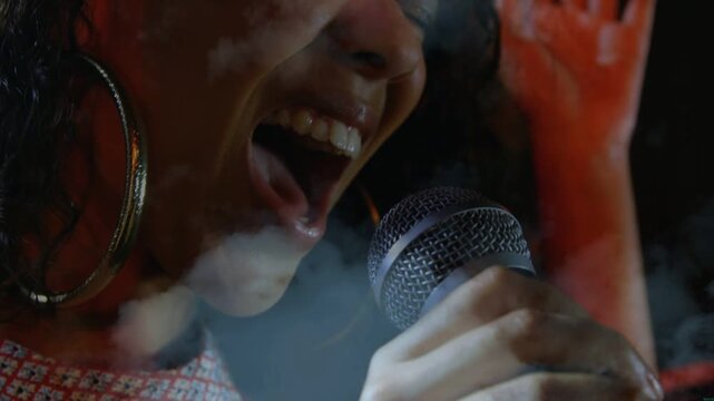 Female singer singing into mic on music stage, rising phrase driving vocal power amid red smoke