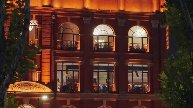 Lit arched windows on brick facade showing warm dinner service inside restaurant staff arranging tables, patrons seated at window tables, balcony seating with ambient lamps, marquee casting amber