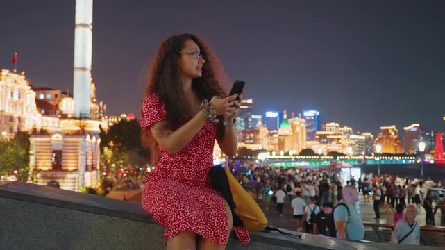 Black woman in red dress on river esplanade at night using phone, illuminated skyline and crowded promenade in background, travel blogger capturing photos and messages, warm city lights and cinematic