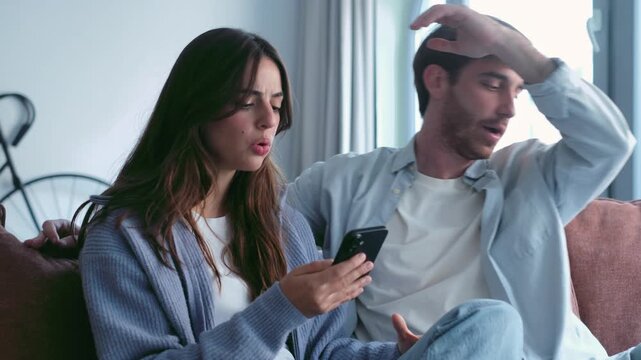A young couple engages in a tense discussion, looking at a smartphone together on the sofa. Their body language suggests a disagreement or problem, highlighting relationship issues in the digital age.