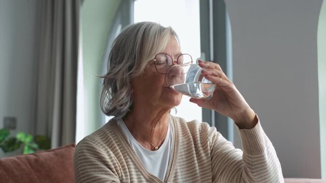 A serene elderly woman with glasses calmly drinks water from a glass, highlighting the importance of hydration and self-care for health and wellness. She is enjoying a quiet moment at home.