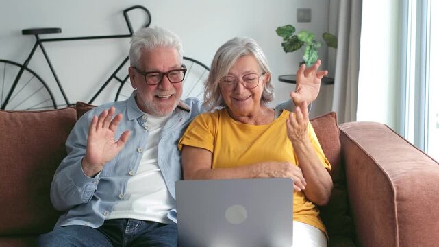 A happy elderly couple waves and smiles during a video call on their laptop, connecting with family or friends from the comfort of their home. They embrace modern technology for remote connections.