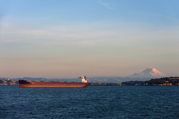 Fototapeta premium Large cargo ship transporting goods in Puget Sound at sunset, international freight, trade, commerce, and economics in action, Mt. Rainier and port of Seattle in background 