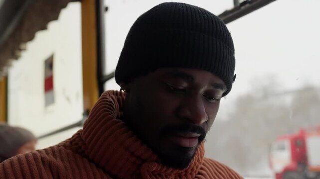 Black man commuter gazing through window, winter light, wool sweater and beanie, candid close up portrait showing thoughtful pensive expression, frames shift from checking ticket to scribbling notes
