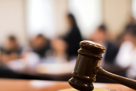 Gavel on a wooden table in a courtroom with blurred people in the background.