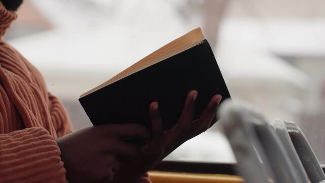 Hands holding book on bus window intimate closeup of reader in knit sweater, soft winter light, snowy city through glass, quiet commute scene, fingers turning pages, focused pace and cozy mood