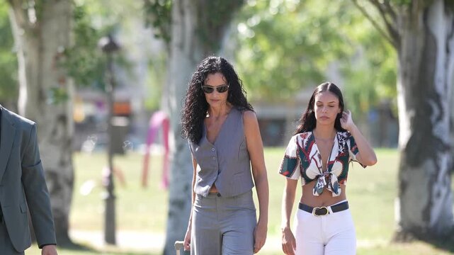 Three people walk through a sunlit park. Man carries a briefcase, steps relaxed. Woman pulls a suitcase, wears sunglasses. Another woman walks beside her, head in hand. Trees frame their path