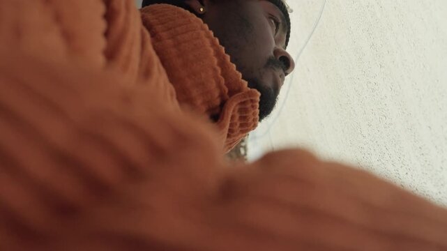 Black man gazing through rainy window, wearing orange knit sweater and black beanie, leaning against train glass with droplets, reflective mood of commuter during cold season, muted cinematic tones,