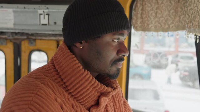 Black man inside yellow city bus, orange knit sweater and black beanie visible, hands near mouth to warm, gaze on snowy street, alternating roles across frames as driver, onboard conductor,