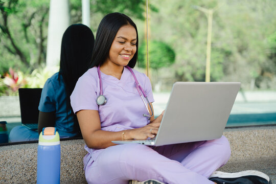 Latin hispanic healthcare worker wearing scrubs and stethoscope, studying and working on laptop while sitting outdoors